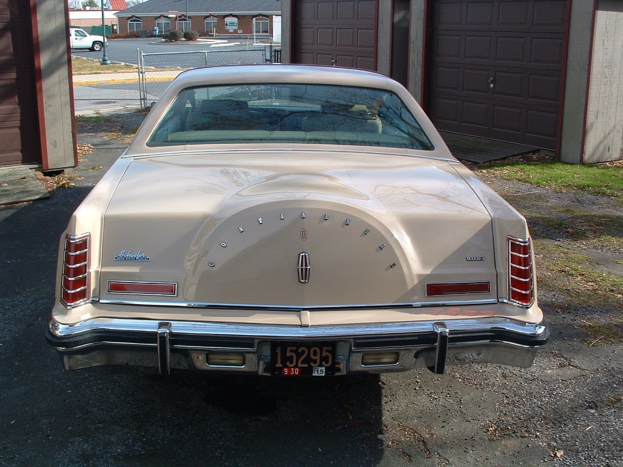 Rear view of a vintage beige Oldsmobile Cutlass with a spare tire cover, parked on a gravel driveway with buildings and a parking lot in the background.