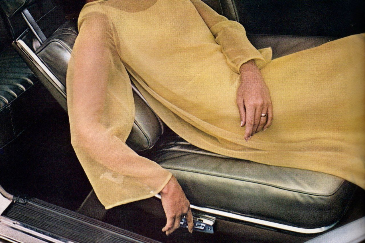 A person lying on their side in the back seat of a car, with their hand resting on their abdomen. They are wearing a light yellow dress and a ring on their finger.