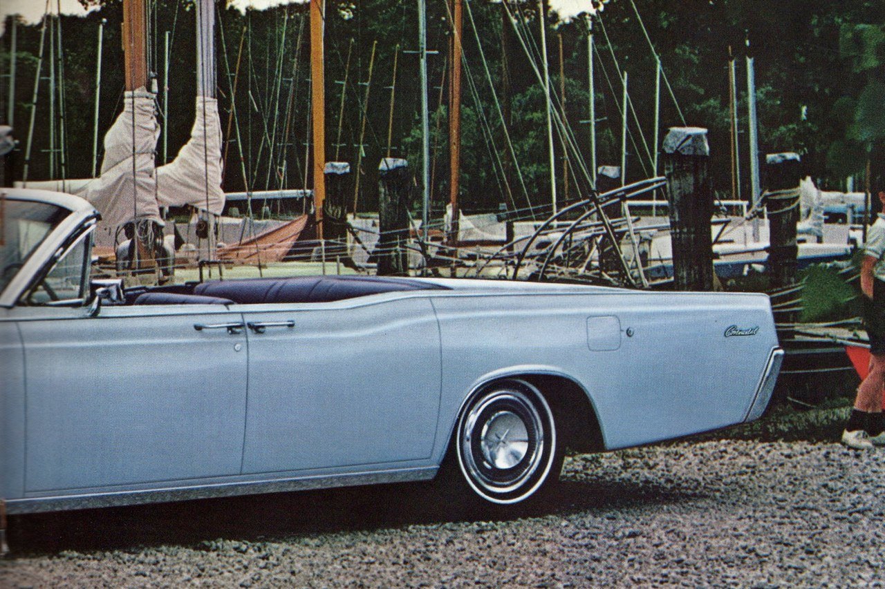 A vintage light blue Chevrolet Caprice convertible parked near sailboats at a marina with trees in the background.