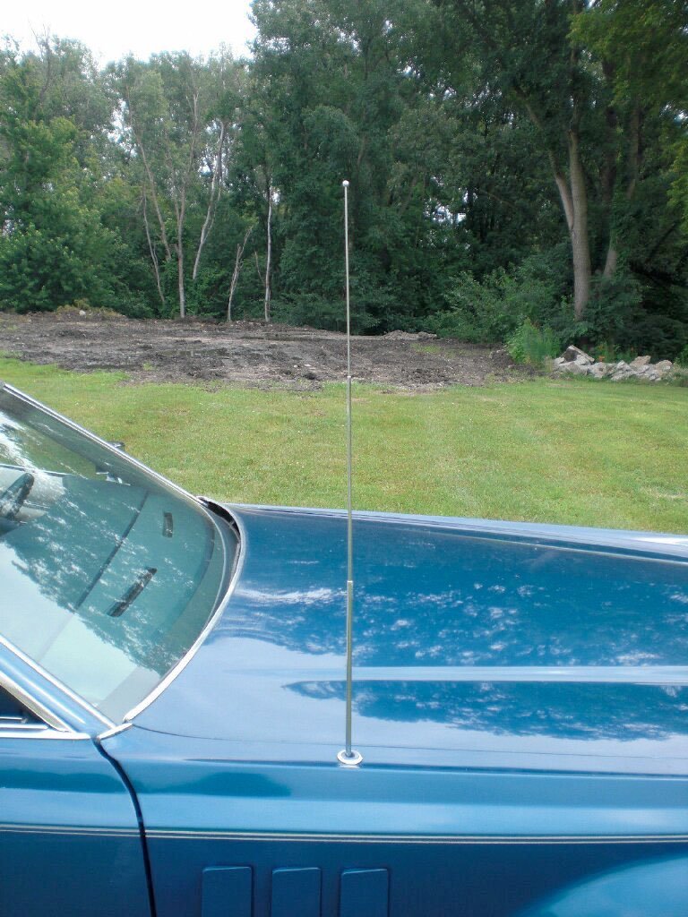 Close-up of a vintage blue car with an extended radio antenna on its hood, parked outdoors with green grass and trees in the background.
