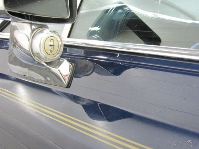 Close-up of a vintage car's front fender with a shiny chrome bumper and a round headlight, parked on a street.