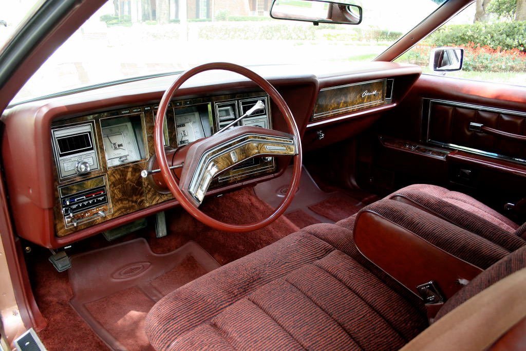 Interior of a vintage car with a dashboard, steering wheel, and front bench seat, featuring wood accents and red upholstery.