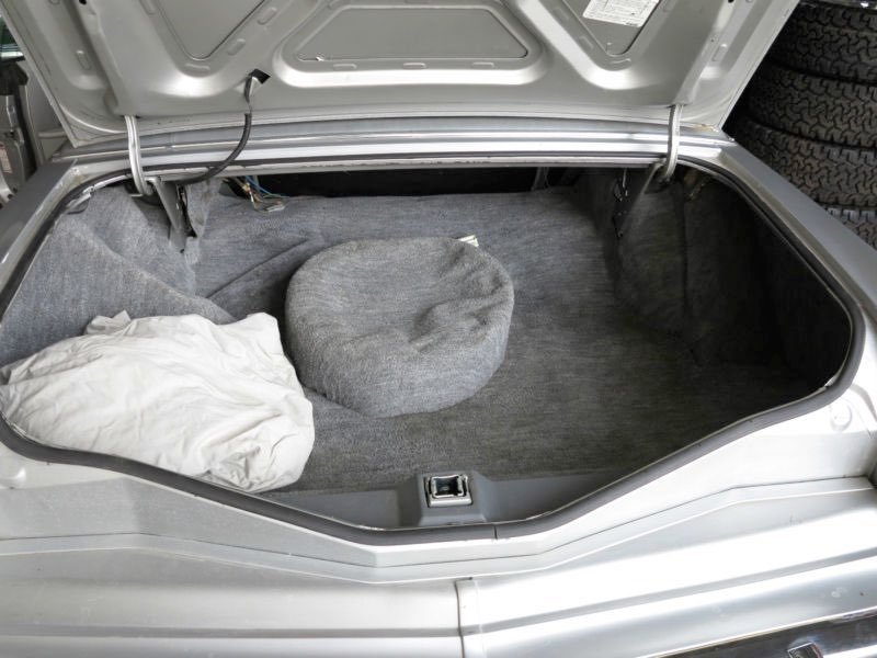 The trunk of a silver car with gray carpeting, containing a white cloth item and a round gray cushion or pillow.