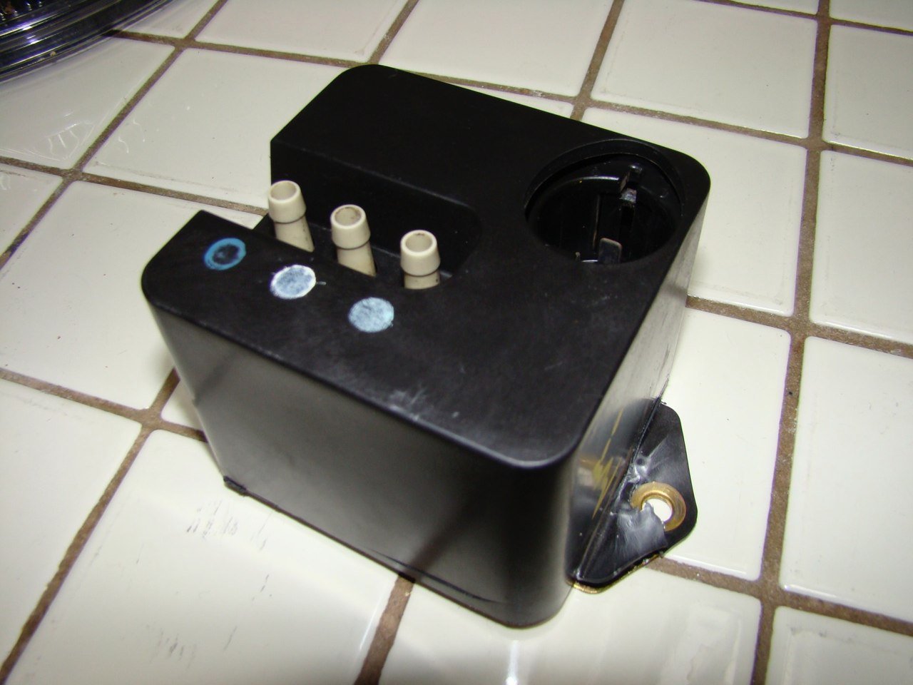 Black power strip with three white outlets and various holes on a white tiled surface.
