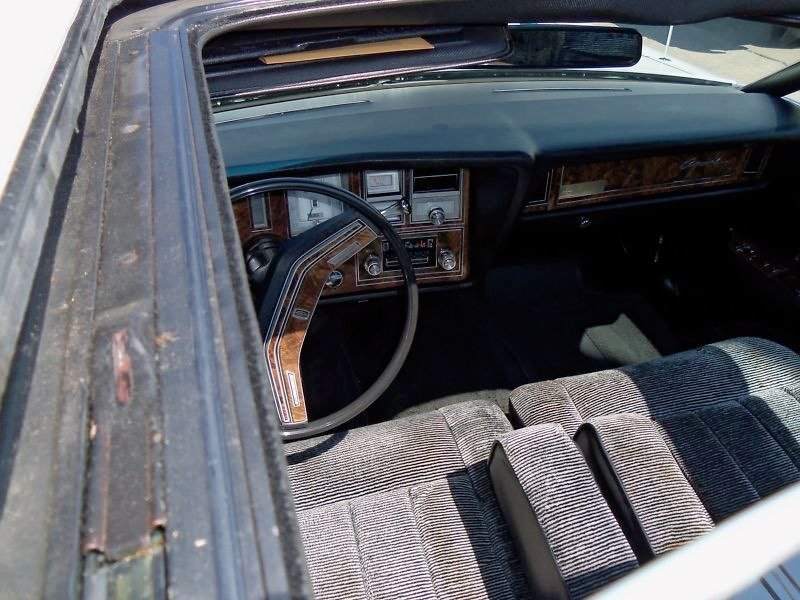 Interior of a vintage car showing steering wheel, dashboard with wood paneling, front bench seat with gray and black striped fabric.