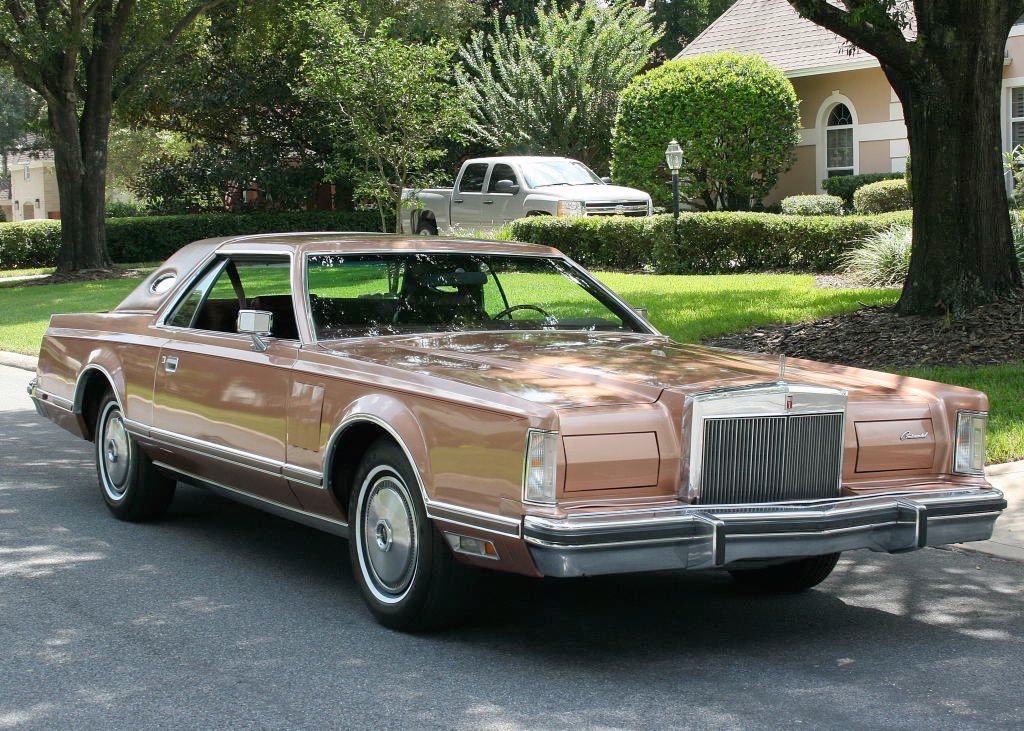 A vintage Lincoln Continental luxury car parked on a residential street with trees and a house in the background.