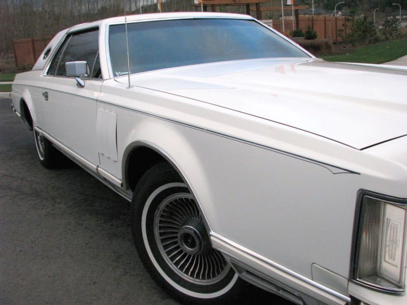 A vintage white luxury car parked outdoors with a wooden fence and trees in the background.