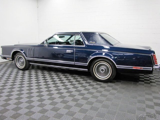 A classic dark blue luxury car with chrome details parked indoors on a checkered tile floor with a white wall background.