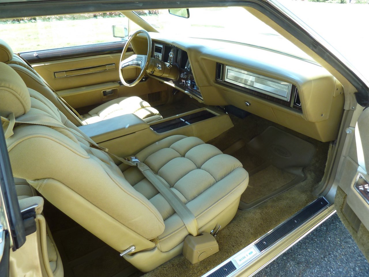 Interior of a vintage car, featuring beige seats, dashboard, and steering wheel, with a large windshield and side window showing the outdoors.