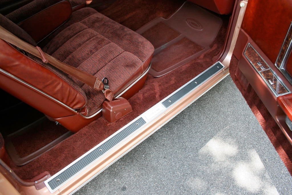 The interior of a vintage car with brown and red upholstery, including a front seat, floor mats, and a metal door sill with a black rubber strip, along with a dark carpeted floor.