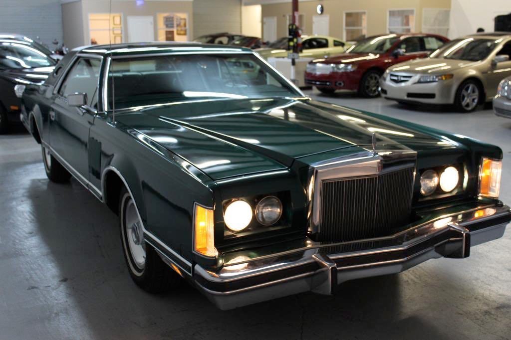 A vintage green luxury car with bright headlights parked in an indoor showroom with other modern cars in the background.