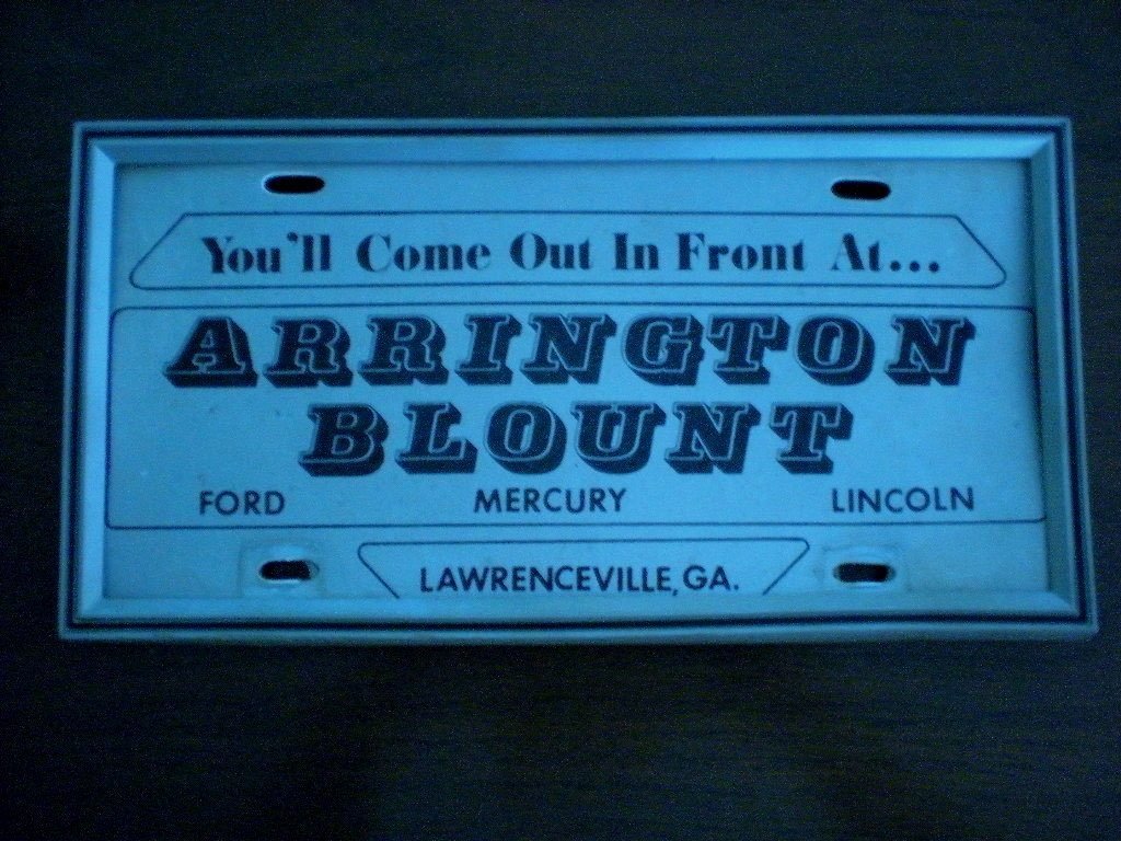 A vintage license plate from Lawrenceville, Georgia, with the text 'You'll Come Out In Front At... ARRINGTON BLOUNT' and the brands Ford, Mercury, and Lincoln.