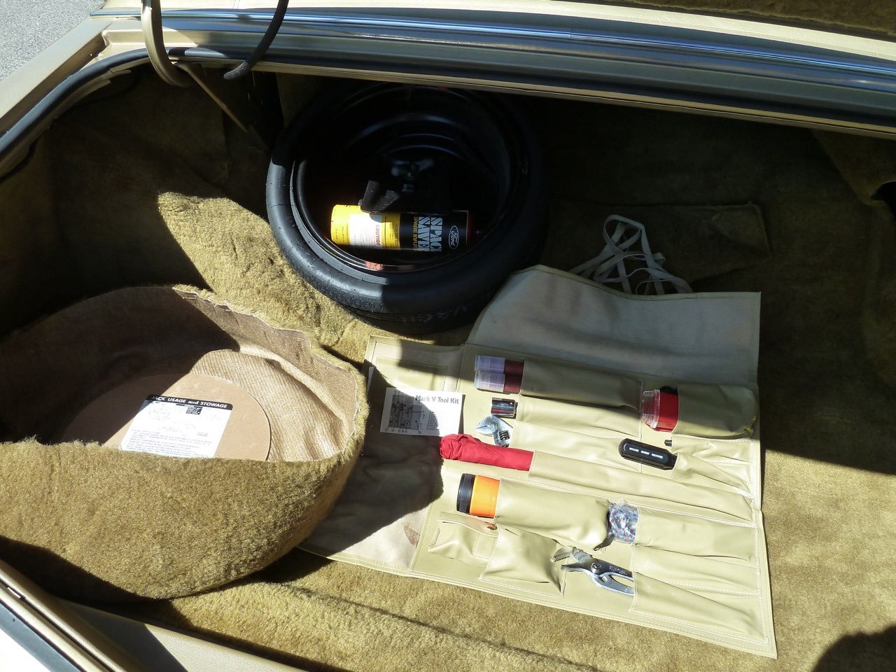 Car trunk containing a spare tire with a bottle, a beige tool roll with various tools and small bottles, and a coiled white extension cord.