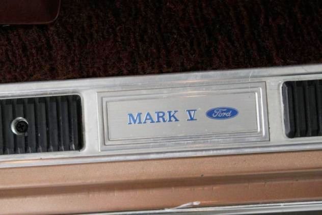 Close-up of a Ford car trunk badge that reads 'Mark V' with Ford logo, surrounded by black vent-like details.