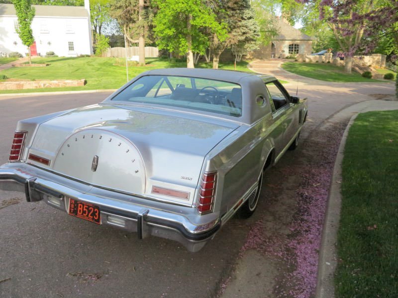 A vintage silver car parked on the side of a street in a residential neighborhood with green lawns and trees.