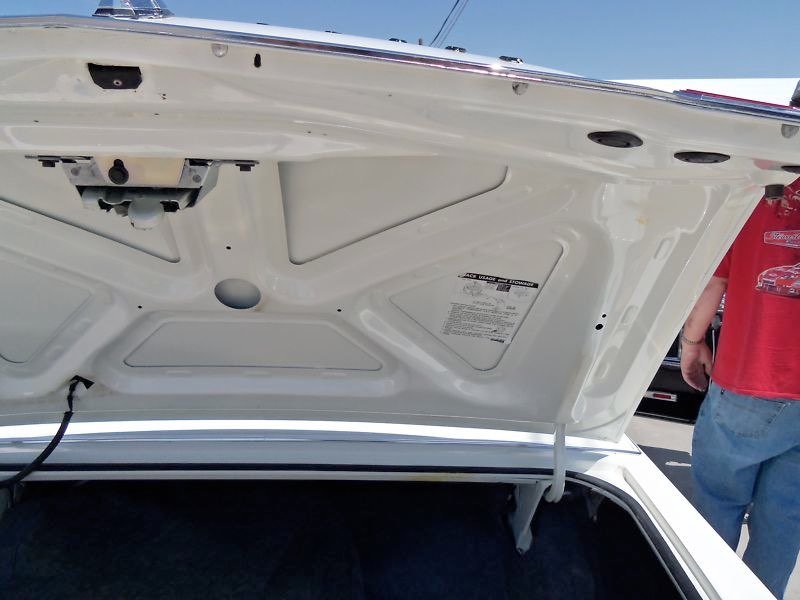 The image shows the open trunk of a white car with the underside of the trunk lid visible, and part of a person wearing a red shirt and blue jeans standing nearby.