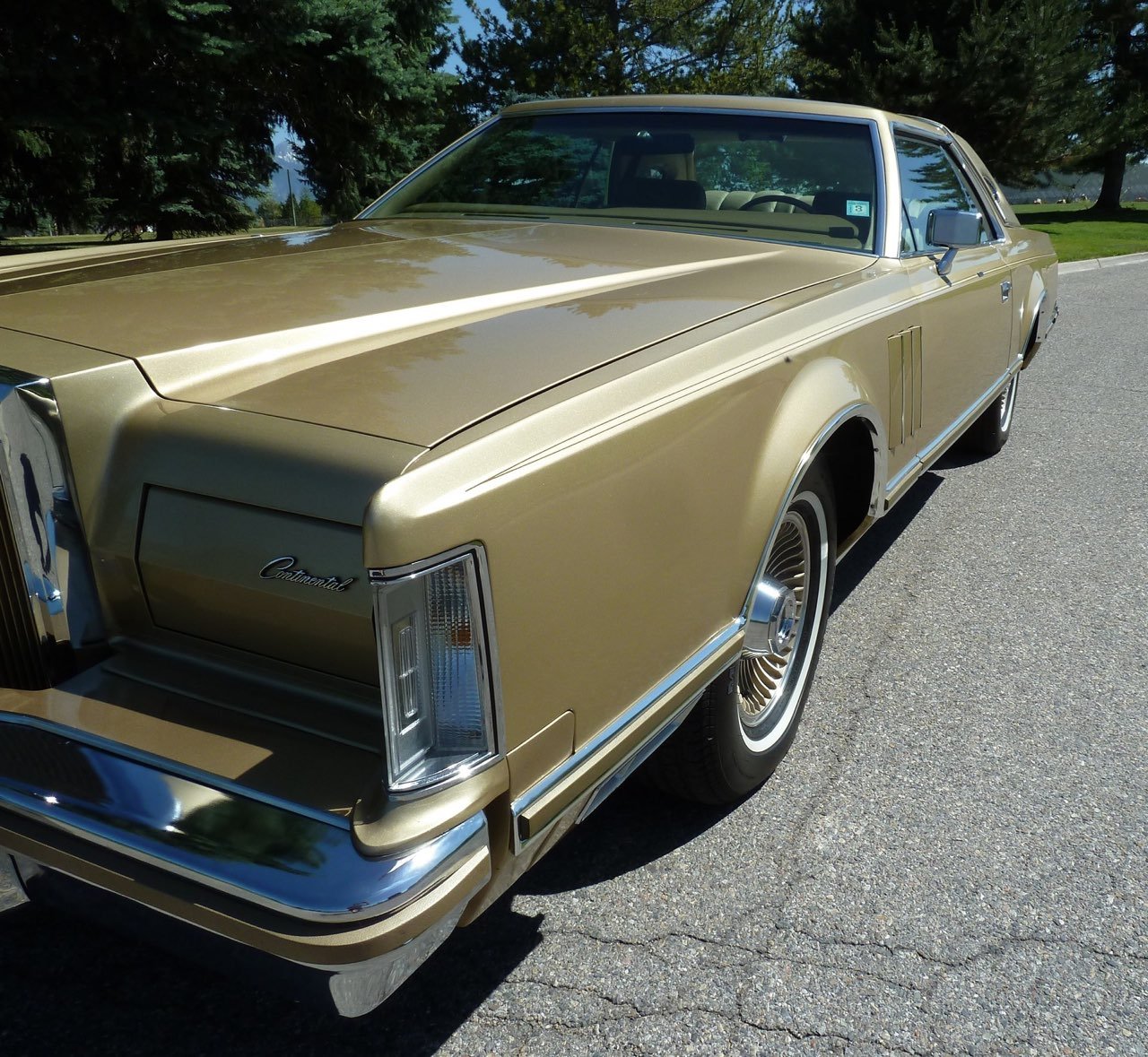 A vintage gold-colored luxury car parked on a street with green trees in the background.