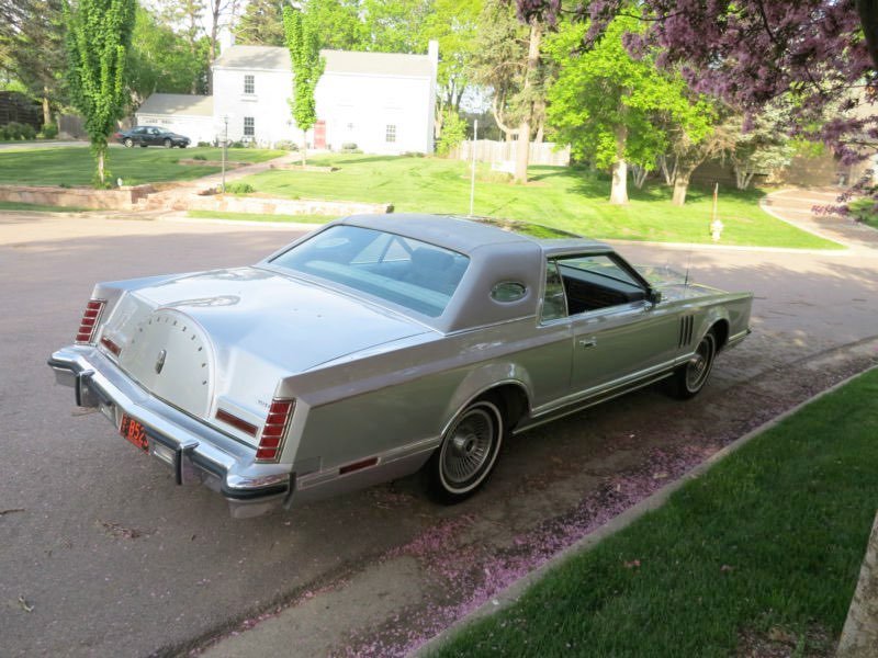 A vintage silver coupe parked on a suburban street with green lawns and trees in the background.
