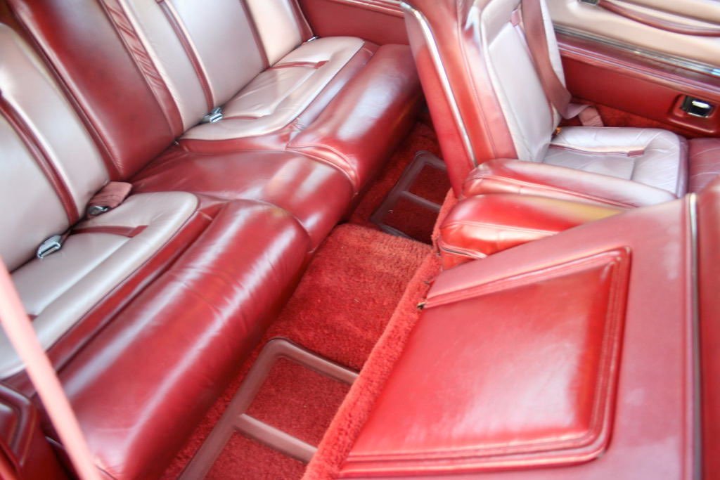 Red and beige leather interior of a vintage car, showing the back seat, front seats, and red carpet.