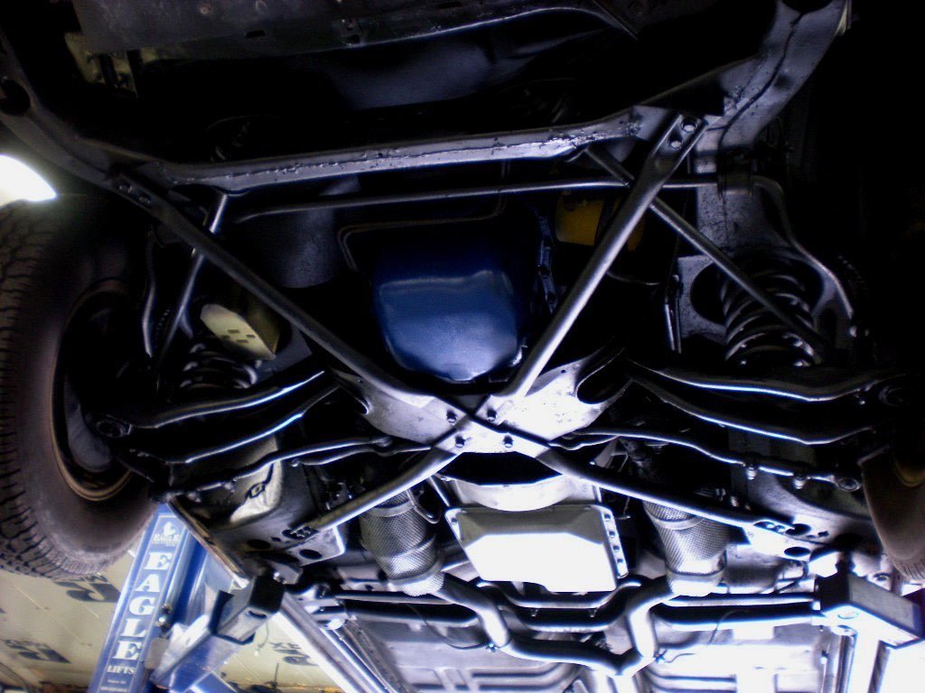 Underneath view of a car with visible suspension, exhaust system, and a roll cage, taken in a garage or workshop.