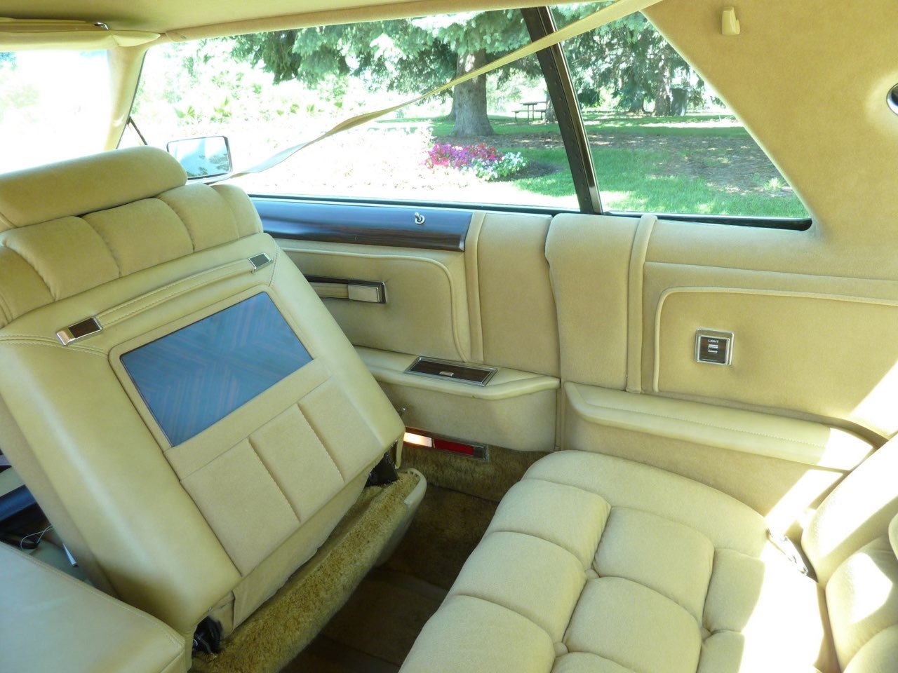 Interior of a vintage car with yellow leather seats, a screen on the back of the front seat, and a window view of trees and flowers outside.