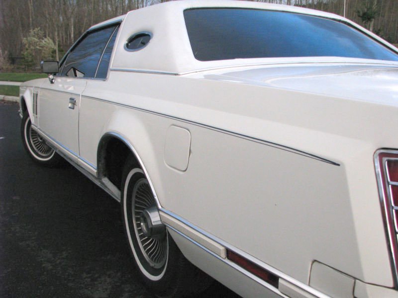 A white vintage car with a distinctive rear design, parked outdoors on a paved surface with trees in the background.