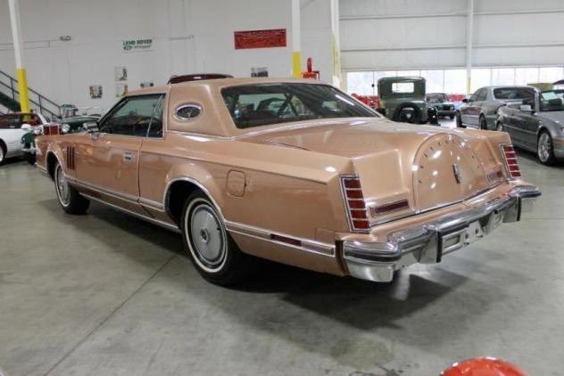 A classic beige luxury sedan with a roof extension, parked in an indoor car showroom, surrounded by other vintage cars.