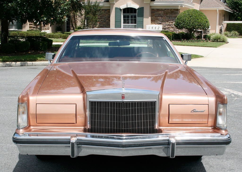 A vintage copper-colored Cadillac car parked on a residential street in front of a house with landscaped lawn and trees.