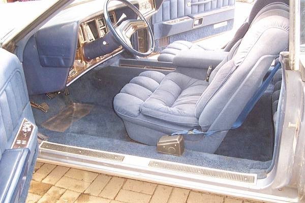 Inside of a vintage car showing a plush, light blue driver's seat, dashboard, and steering wheel with a black cover.