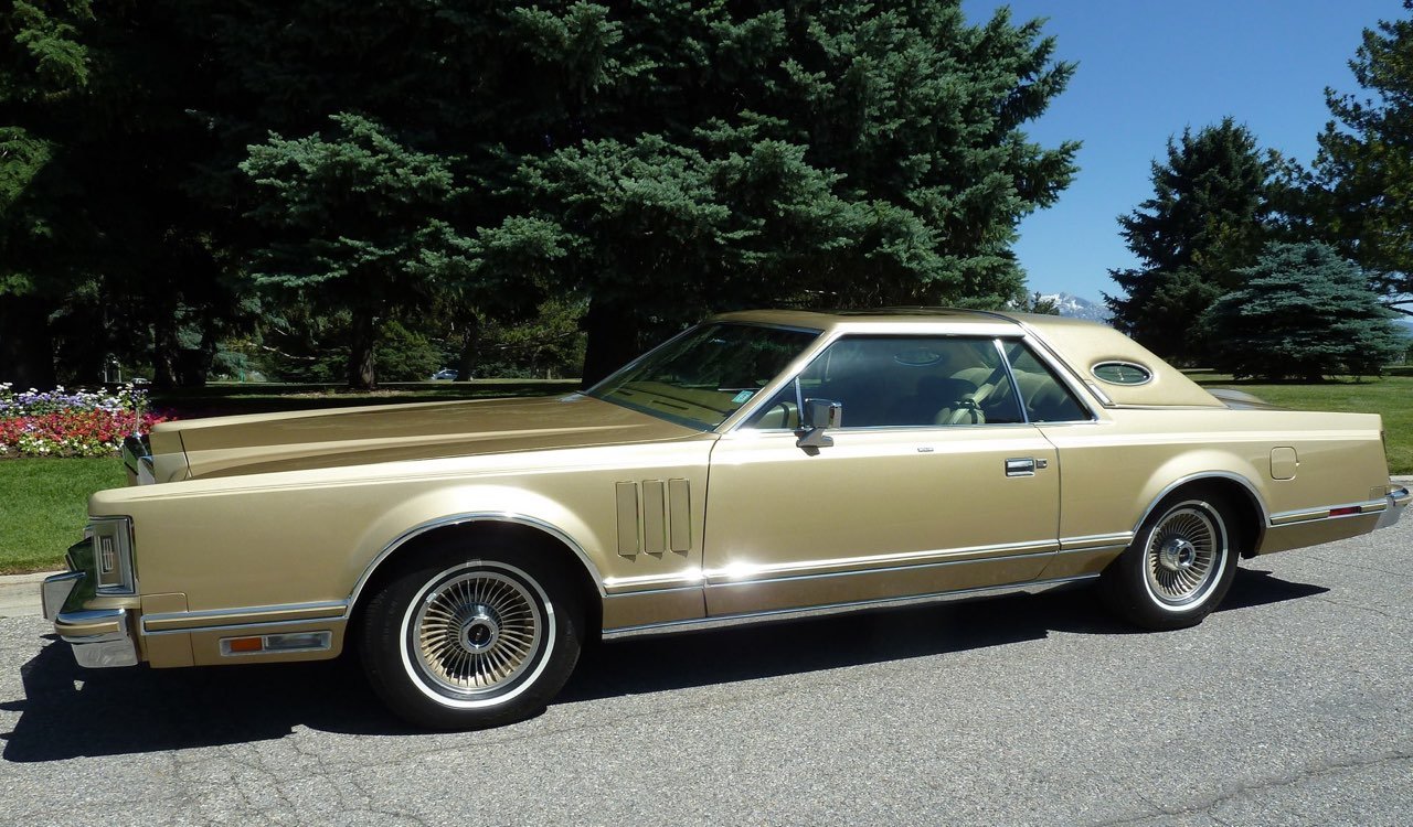 A vintage gold-colored luxury car parked on a paved road with green trees and a yard with flowers in the background.