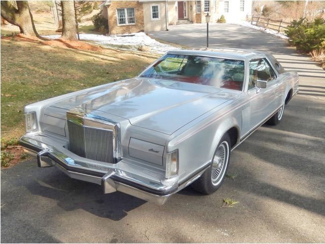 Silver vintage luxury car parked on driveway in front of house with trees and a lamp post nearby.