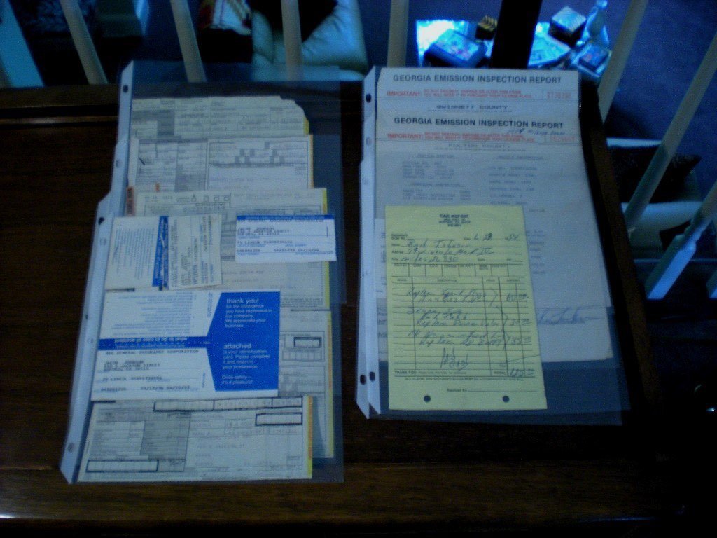 A table with multiple vehicle-related documents, including Georgia emission inspection reports, receipts, and paperwork, under plastic sleeves. The background shows a chair and part of a person.