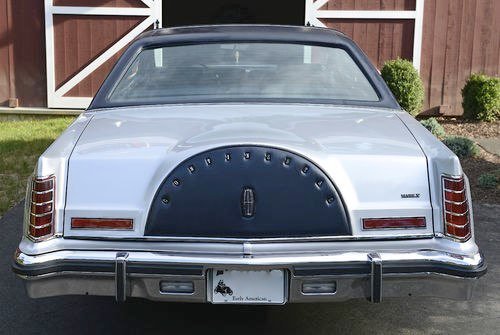 Rear view of a vintage silver car with a full-sized spare tire mounted on the trunk, parked in front of a wooden fence with small green bushes.