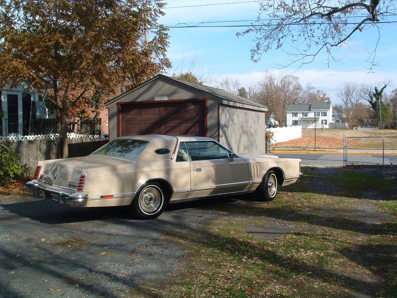 A vintage beige car parked in a driveway next to a small garage with a brown door, surrounded by trees and suburban houses under a partly cloudy sky.