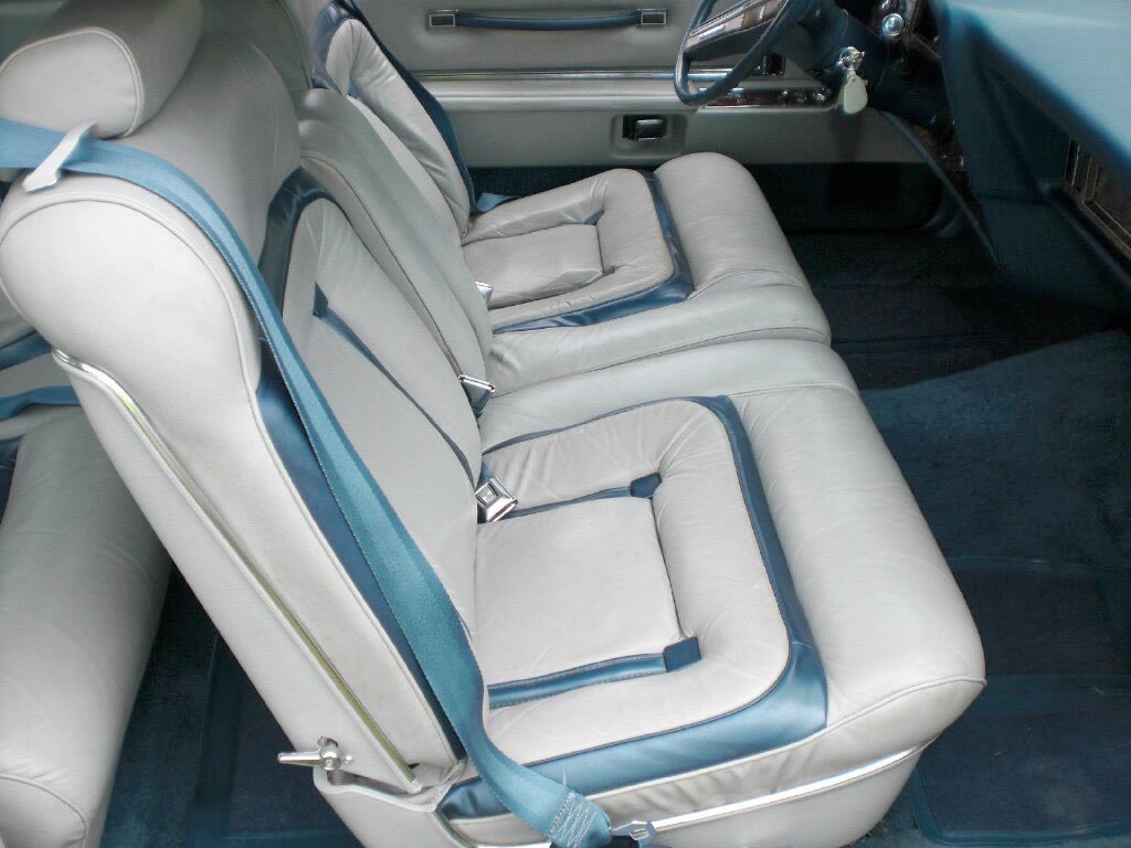 Interior of a vintage car showing gray leather seats with blue trim and seat belts, dashboard, and steering wheel.