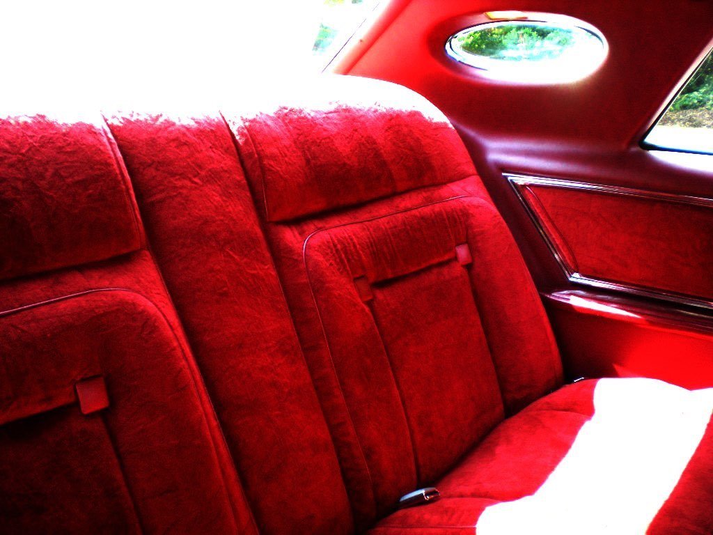 Red velvet interior of a vintage car's back seat area with patterned upholstery, a curved window, and a porthole window on the car's ceiling.