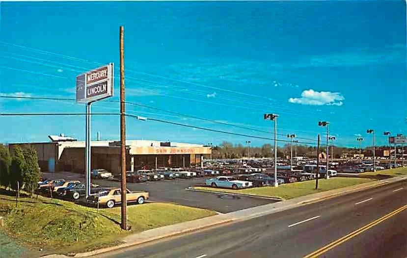 Parking lot outside a building with a sign for 'San Johnson' surrounded by cars, green grass, street with yellow lines, and a blue sky with some clouds.