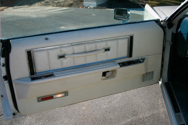 Close-up of a vintage car interior door panel with a white padded armrest, a chrome window handle, and a small storage compartment, set against an outdoor background.