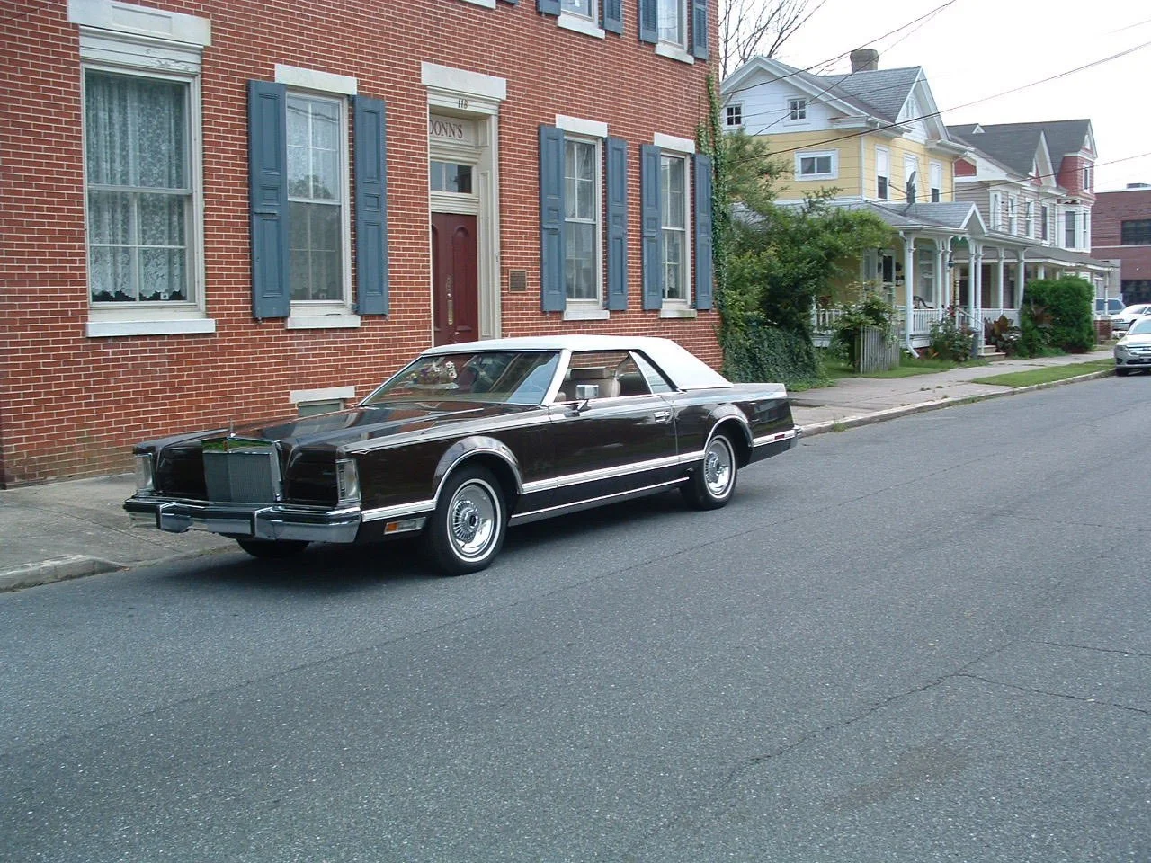 A vintage black car with a white roof parked on a residential street with historic brick and wooden houses