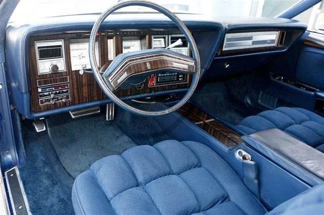 Interior of a vintage car with a wood grain dashboard, a steering wheel with a metallic center console, blue upholstered seats, and an analog radio.