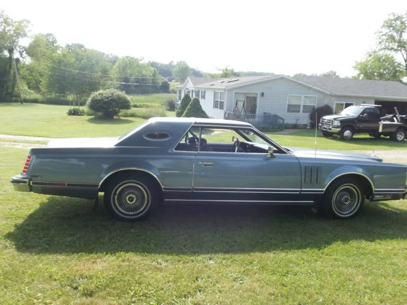 A vintage blue coupe car parked on a lawn in front of a house with a white exterior and a black truck parked nearby.