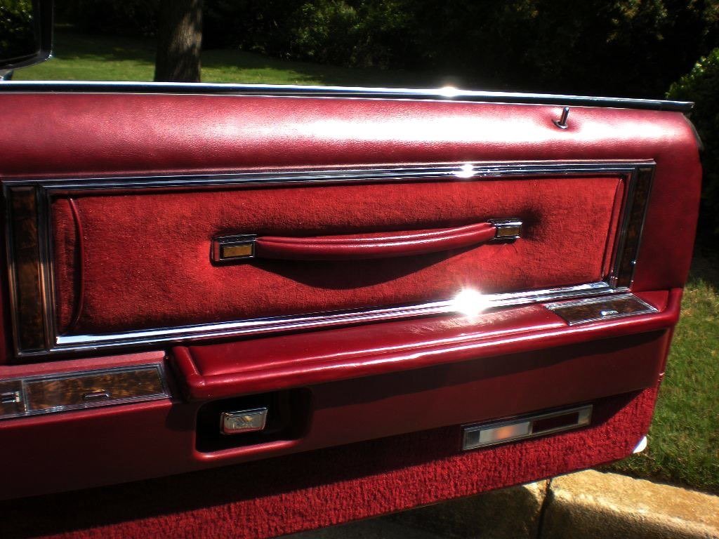 Close-up of a red vintage car door interior with a padded armrest, chrome accents, and a red fabric panel.
