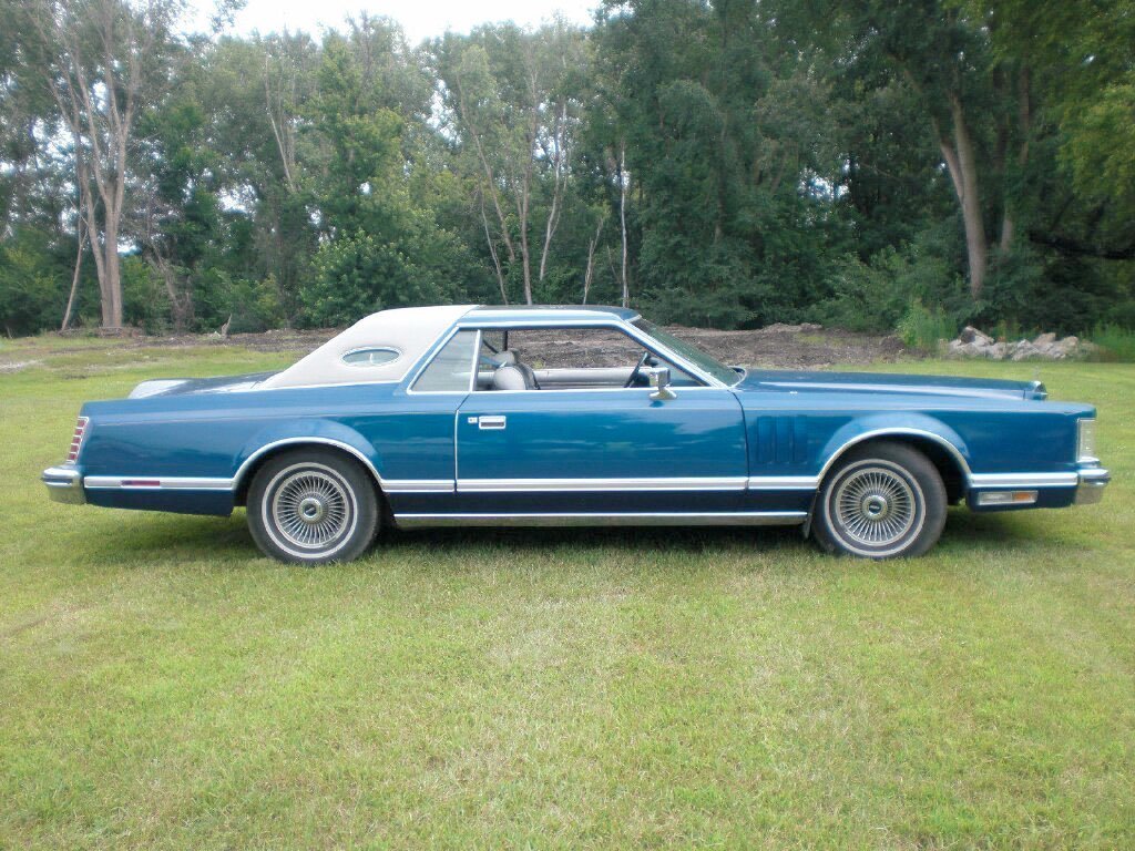 A vintage blue two-door coupe with a white vinyl roof parked on grass, surrounded by trees.