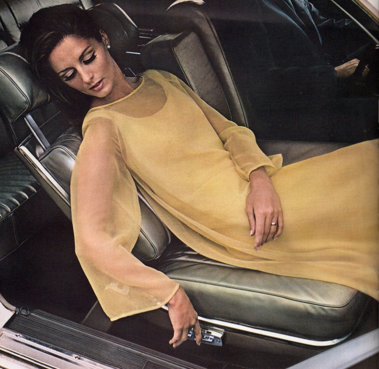 A woman wearing a yellow dress lying on the seats of a car, with her eyes closed and head tilted to the side.