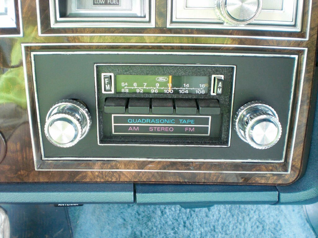 Vintage car stereo with FM/AM radio, quadasonic tape, and stereo controls, set in a woodgrain dashboard.