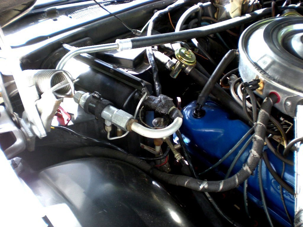 Close-up of a car engine showing various hoses, wires, and metal components including a blue valve cover and a silver air cleaner on top.
