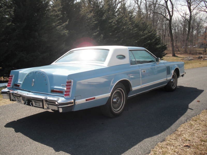 A vintage light blue and white luxury car parked on a paved driveway with trees and grass in the background.