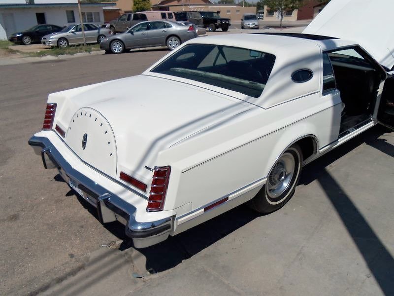 White vintage luxury car with unique circular rear design parked on a street with other vehicles and buildings in the background.