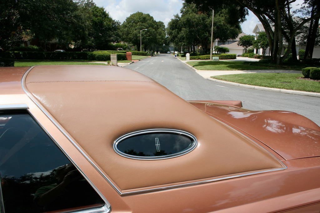 Close-up of a vintage car's tan roof and rear window, parked on a suburban residential street with trees and houses in the background.