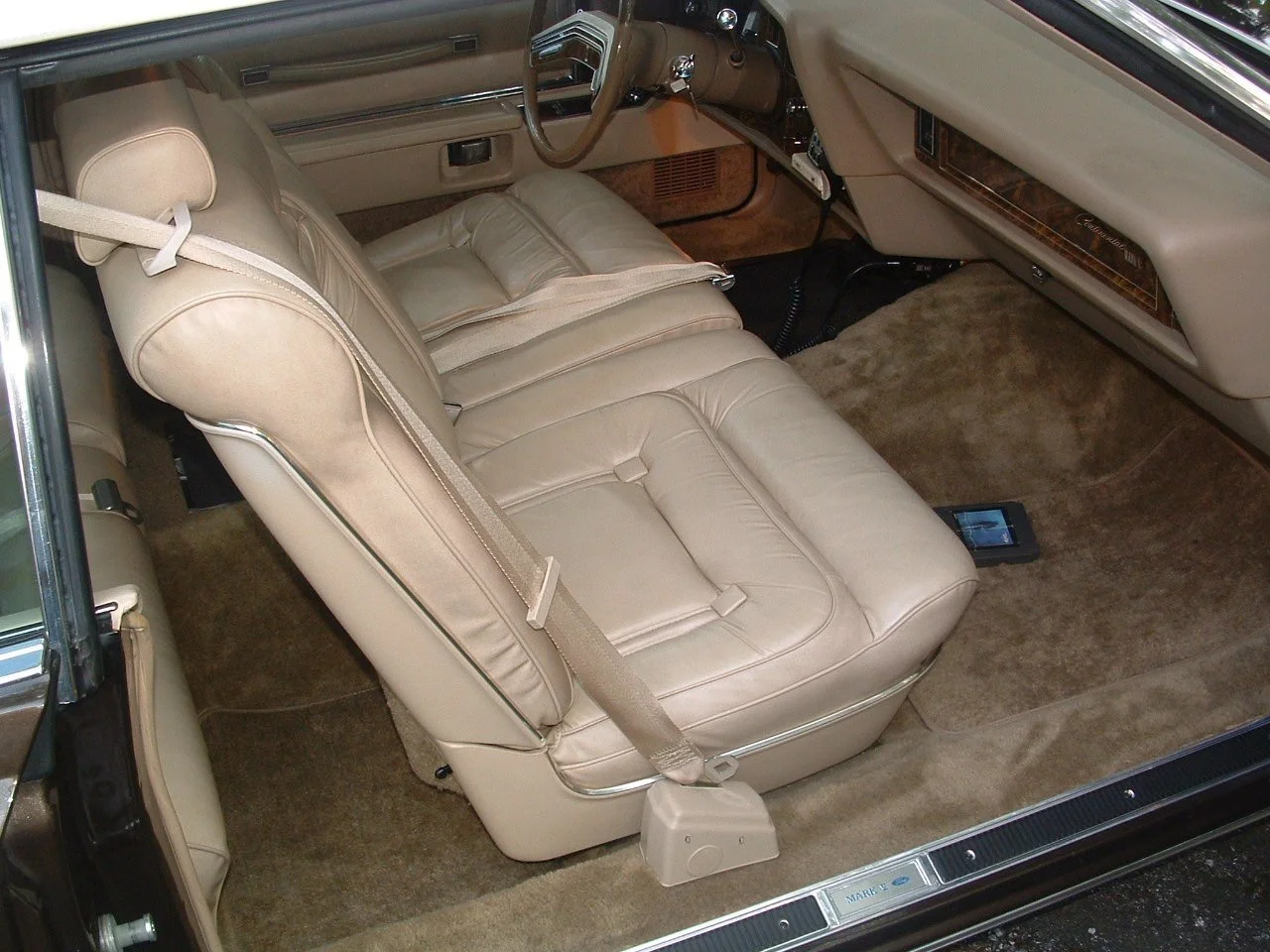 Interior view of a vintage car with cream leather seats, beige carpet, and wood grain dashboard.